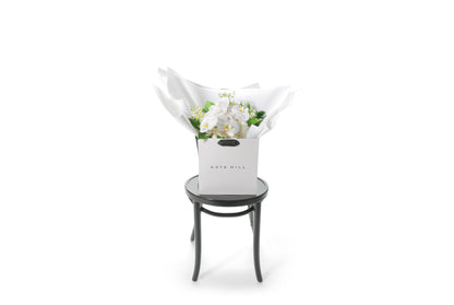 White and green with a pop of yellow lemon medium sized bouquet, beautifully wrapped in white signature presentation. Bouquet is placed into Kate Hill flower bag to ensure the bouquet remains fresh in water. Bouquet bag is sitting on a black bentwood chair with a white background. Wide image of the SASCHA gift bouquet.