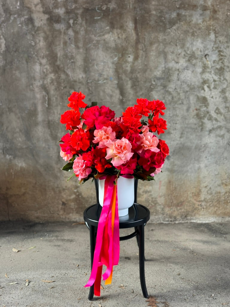 Wide view of the large design full of mixed bright roses. A large white vase displaying masses of orange, pink and watermelon roses which is sitting on a black bentwood chair against a concrete wall.