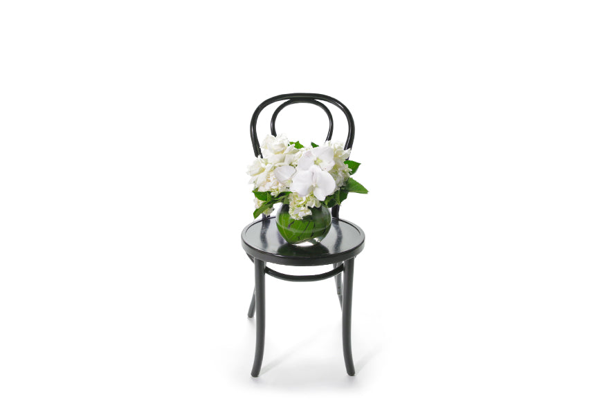 ELISE vase design is classic vase design that displays a balanced mix of white seasonal flowers and green seasonal foliage. Short ball vase, lined with green monsteria leaves and displaying seasonal white flowers and green foliage. Wide image of the vase on chair.