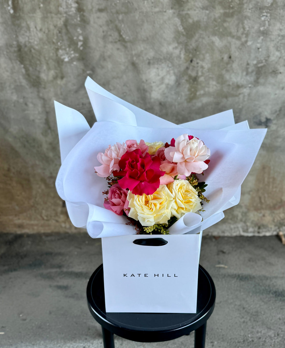 A bouquet of mixed coloured reflexed roses, beautifully wrapped in white and placed into a KHF flower bag. Bouquet sitting on a black bentwood chair against a concrete wall.