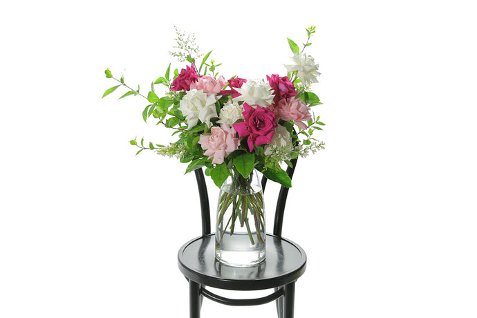 A clear glass tapered vase features 10 stems of mixed coloured roses and green foliage, sitting on a black bentwood chair and white background.