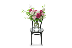 Wide image of Fenella Vase Design. A clear glass tapered vase features 10 stems of mixed coloured roses and green foliage, sitting on a black bentwood chair and white background.