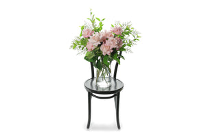 Wide image of Feng pale pink rose vase design. Soft blush pink reflexed roses displayed in a clear glass tapered vase sitting on a black bentwood chair with white background. 