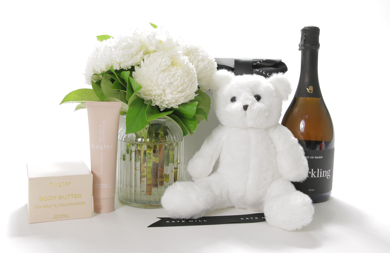 White teddy bear, flowers, and a bottle of sparkling wine on a white surface
