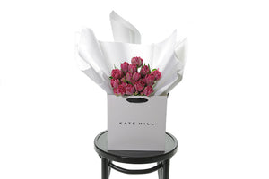 A wrapped bouquet of 20 stems of dark double pink tulips. FRANKIE double Tulip bouquet is beautifully gift wrapped in Kate Hill Flowers signature style that includes layers of premium white and silk paper, finished with Kate Hill signature ribbon. Bouquet is placed into our Kate Hill flower bag. Bouquet bag is sitting on a black bentwood chair.