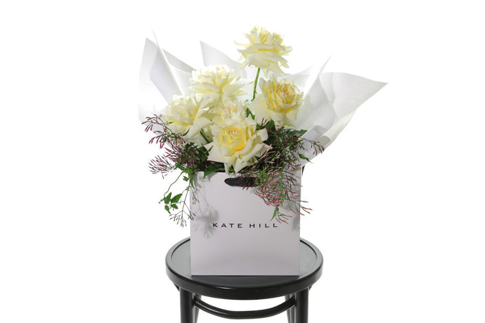 Bouquet of lemon butter roses and seasonal foliage, beautifully wrapped in white signature wrapping. Bouquet displayed in kate hill flower bag, sitting on black bentwood chair with white background.