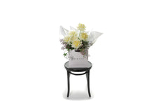 Wide image of GIGI butter bouquet. Bouquet of lemon butter roses and seasonal foliage, beautifully wrapped in white signature wrapping. Bouquet displayed in kate hill flower bag, sitting on black bentwood chair with white background.