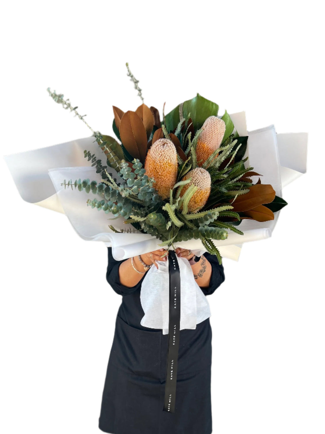 A florist wearing black holding a large graduation bouquet displaying australian native flowers and foliage. Florist standing in front of white wall.