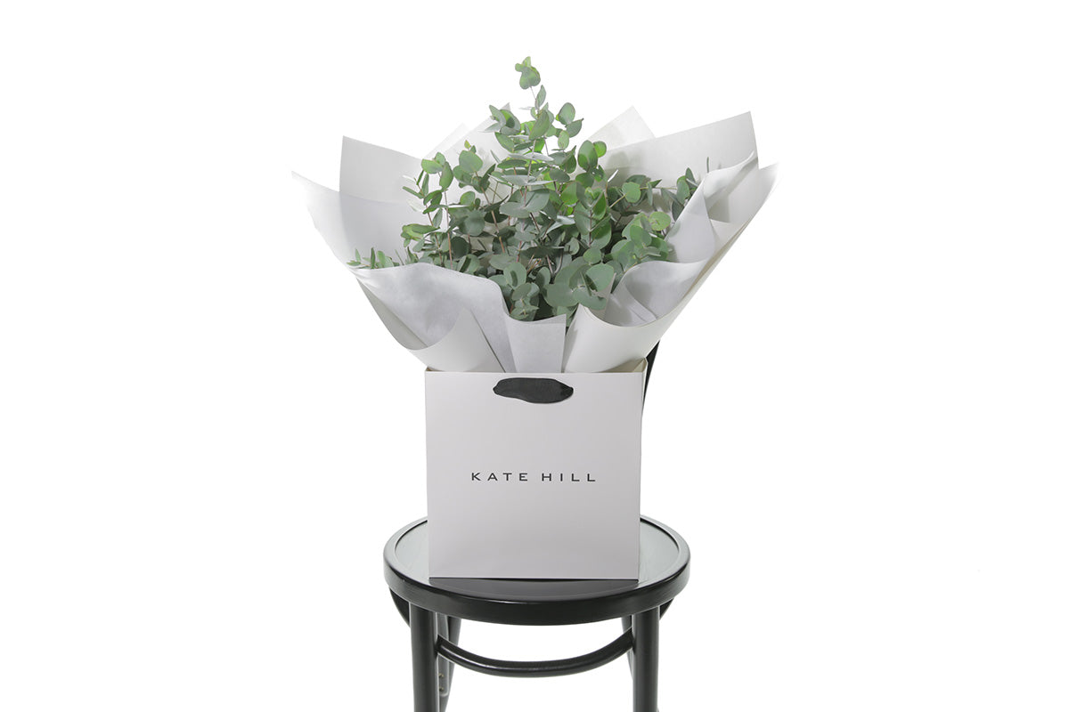 A bouquet of native eucalyptus foliage presented in our signature white wrapping. Flower bag bouquet sitting on black bentwood chair with white background.