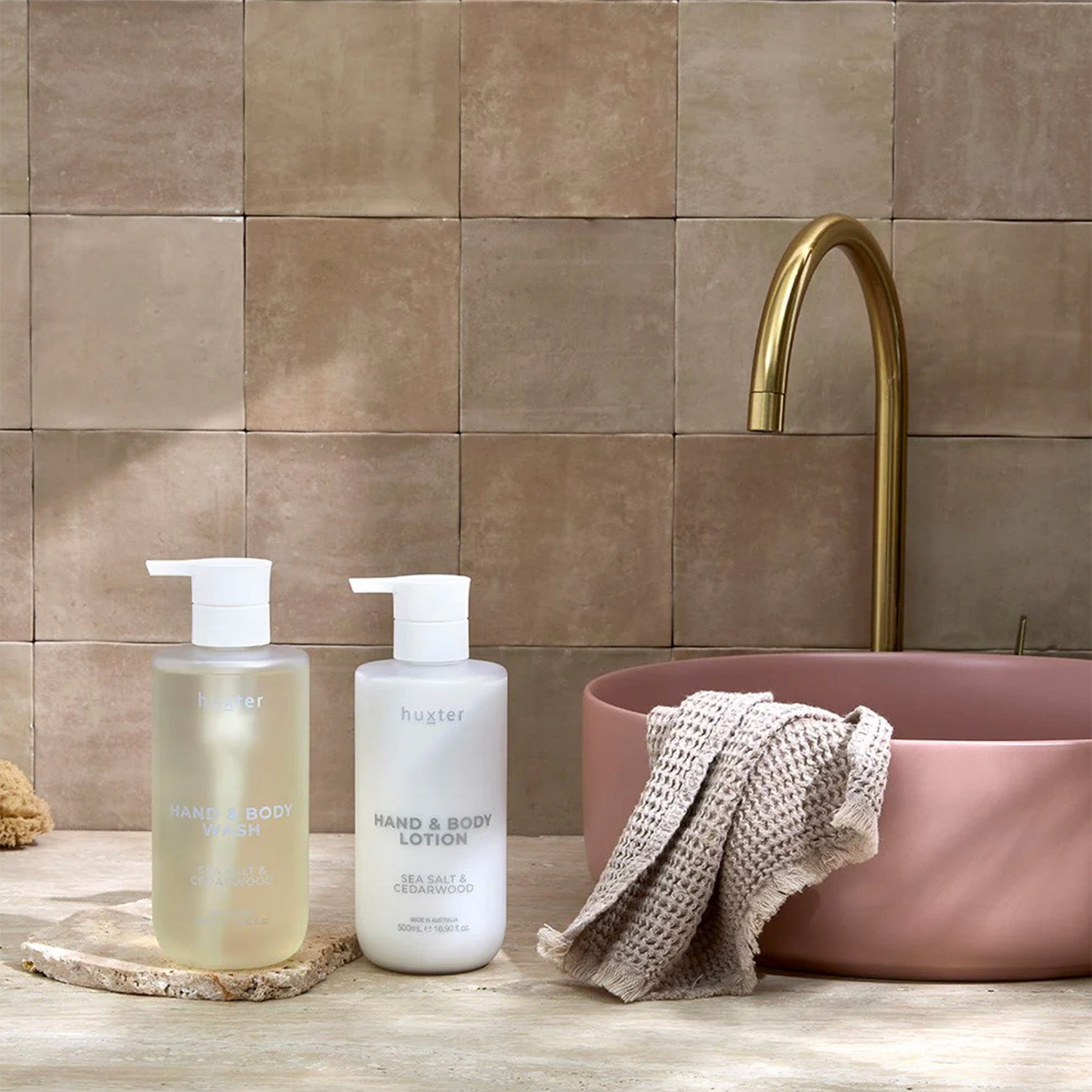 Two bottles of hand and body lotion on a wooden surface with a pink sink and gold faucet in the background.