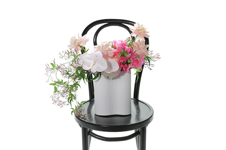 White vase ceramic vase, displaying mixed pink flowers and seasonal foliage. Vase design is sitting on a black bentwood chair with white background.