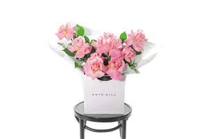 HERMOSA Rose Bouquet is our best selling watermelon pink rose bouquet, curated with Reflexed watermelon pink roses and green seasonal foliage. Large bouquet of watermelon roses presented in our luxury flower bag so it stays fresh in water.  Bouquet bag sitting on a black bentwood chair.