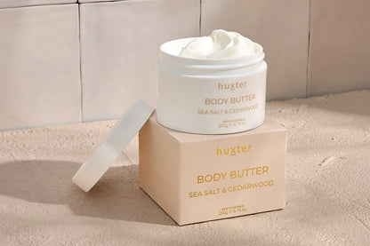 Huxster body butter jar and box on a beige bathroom setting background