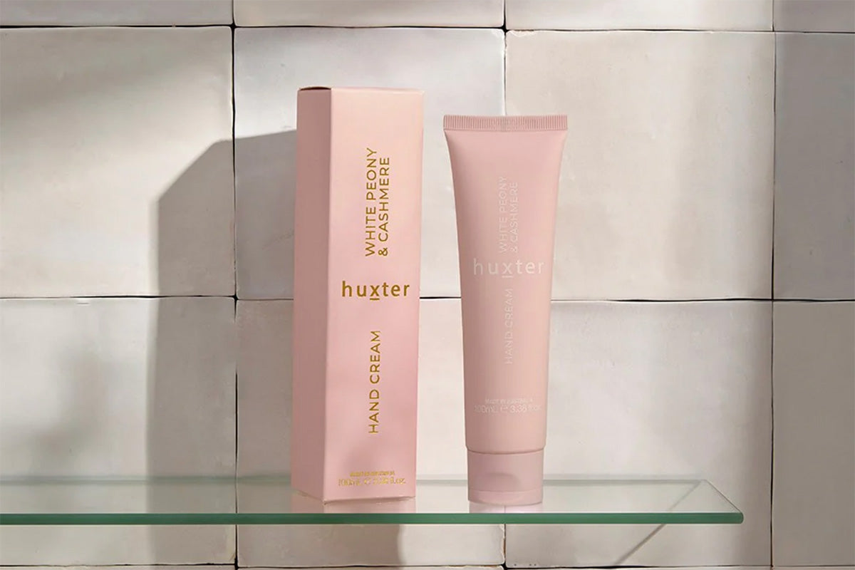 Pink Huxster hand cream and packaging on a glass shelf with a tiled wall background