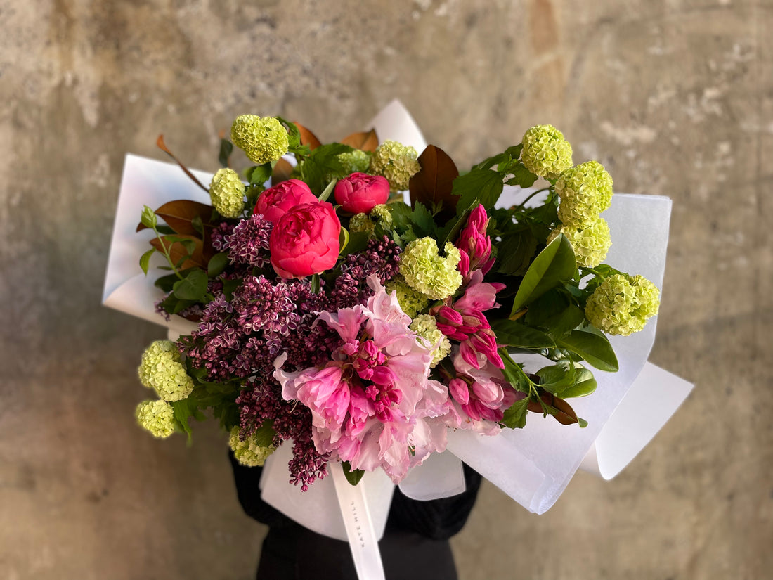 A large mixed jewel tone spring bouquet, wrapped in white signature wrapping. Large Bouquet held in front of concrete wall. 