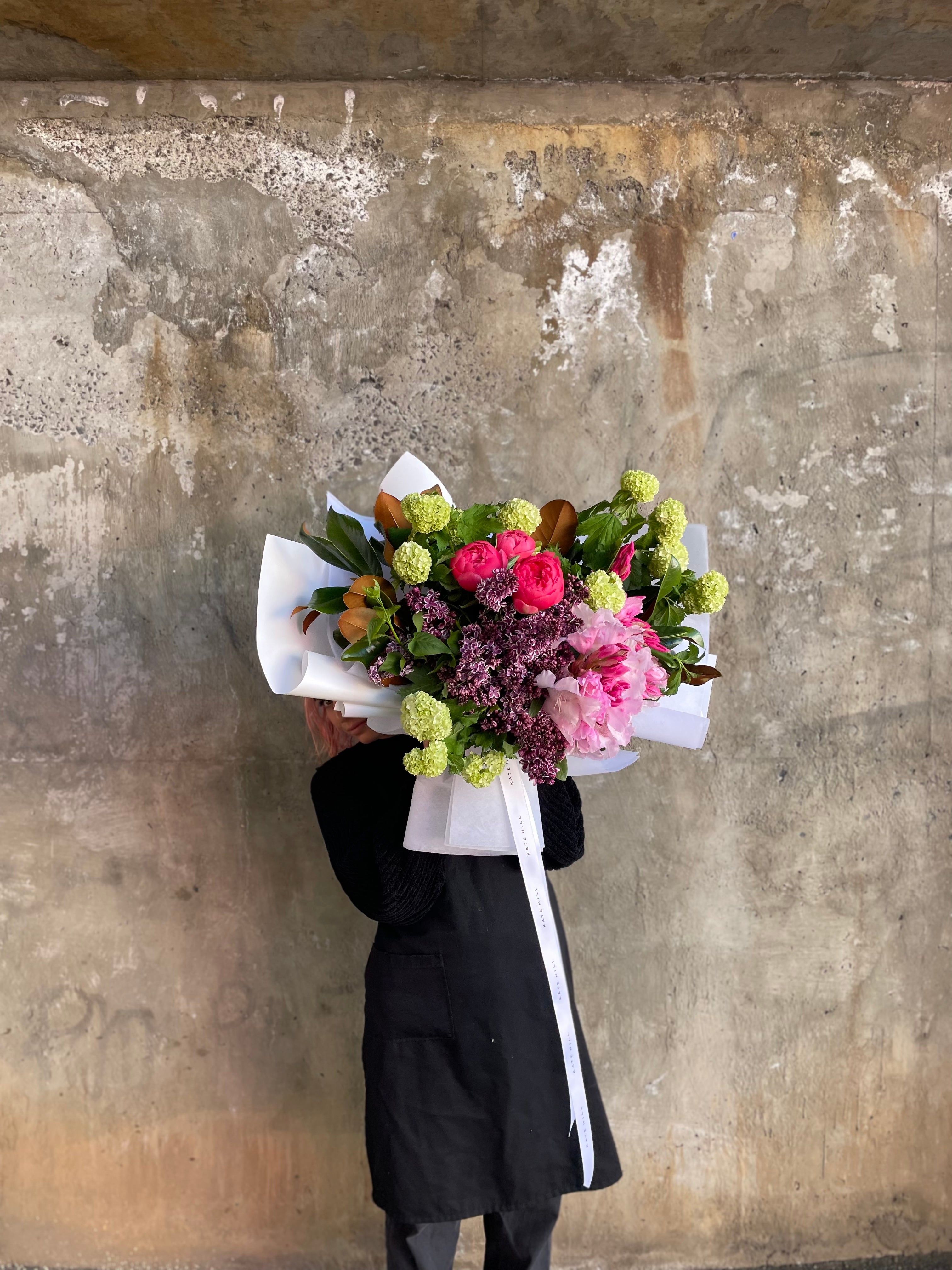 Wide image of bouquet to give an idea of size. A large mixed jewel tone spring bouquet, wrapped in white signature wrapping. Large Bouquet held in front of concrete wall. 