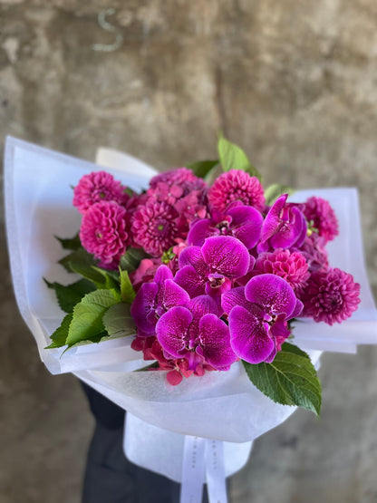 Close up image of magenta bouquet, highlighting the orchids. A bouquet of mixed magenta seasonal flowers such as dahlias, phalaenopsis and raspberry hydrangeas which are beautifully gift wrapped in Kate Hill Flowers signature white wrapping. Florist standing in front of a natural concrete wall holding the bouquet.