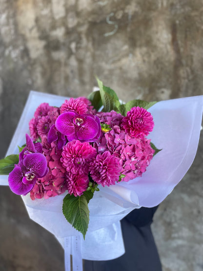 Side angle of the magenta bouquet against the concrete wall. A bouquet of mixed magenta seasonal flowers such as dahlias, phalaenopsis and raspberry hydrangeas which are beautifully gift wrapped in Kate Hill Flowers signature white wrapping. Florist standing in front of a natural concrete wall holding the bouquet.