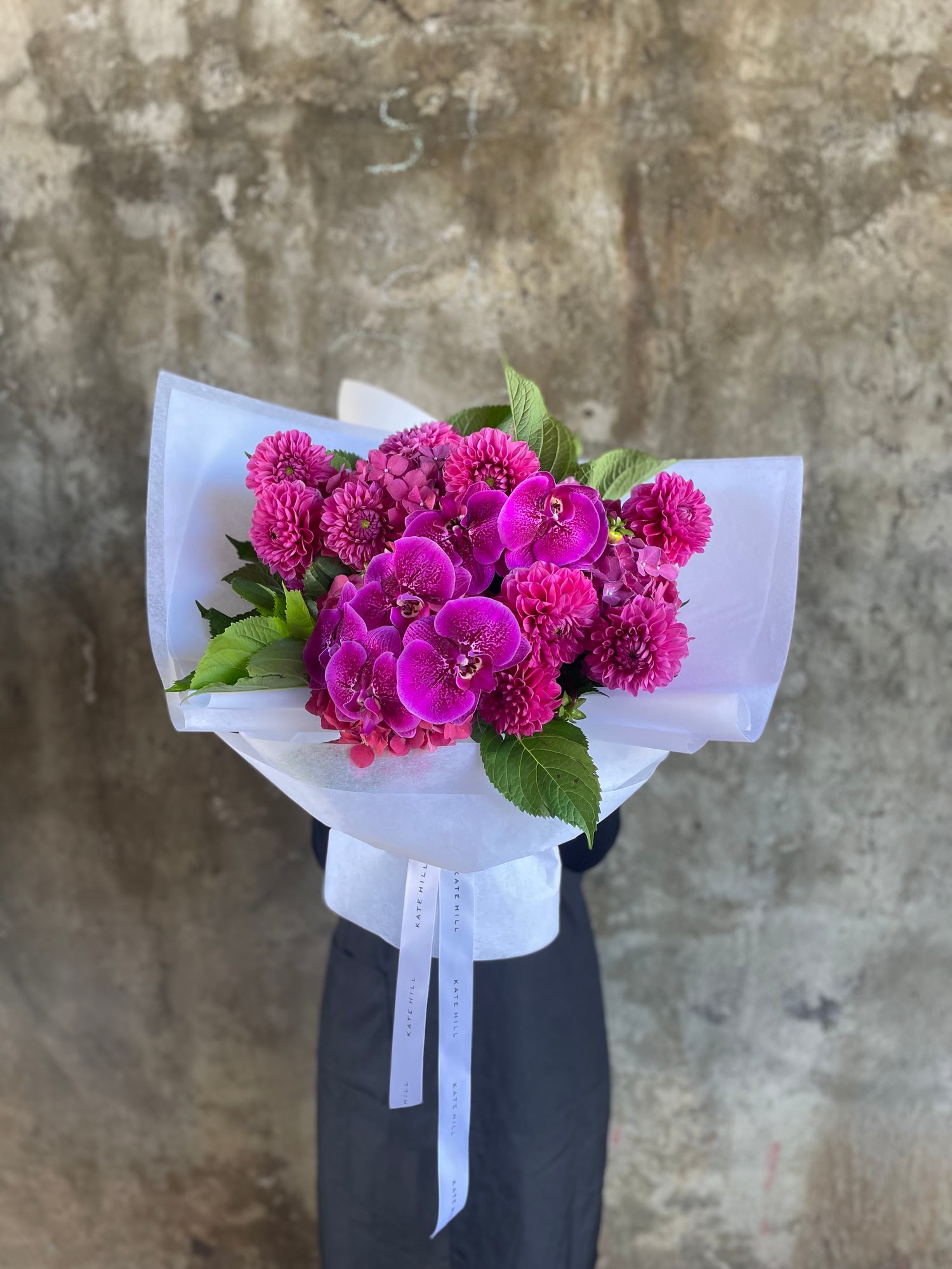 Wide shot of the magenta bouquet. A bouquet of mixed magenta seasonal flowers such as dahlias, phalaenopsis and raspberry hydrangeas which are beautifully gift wrapped in Kate Hill Flowers signature white wrapping. Florist standing in front of a natural concrete wall holding the bouquet.