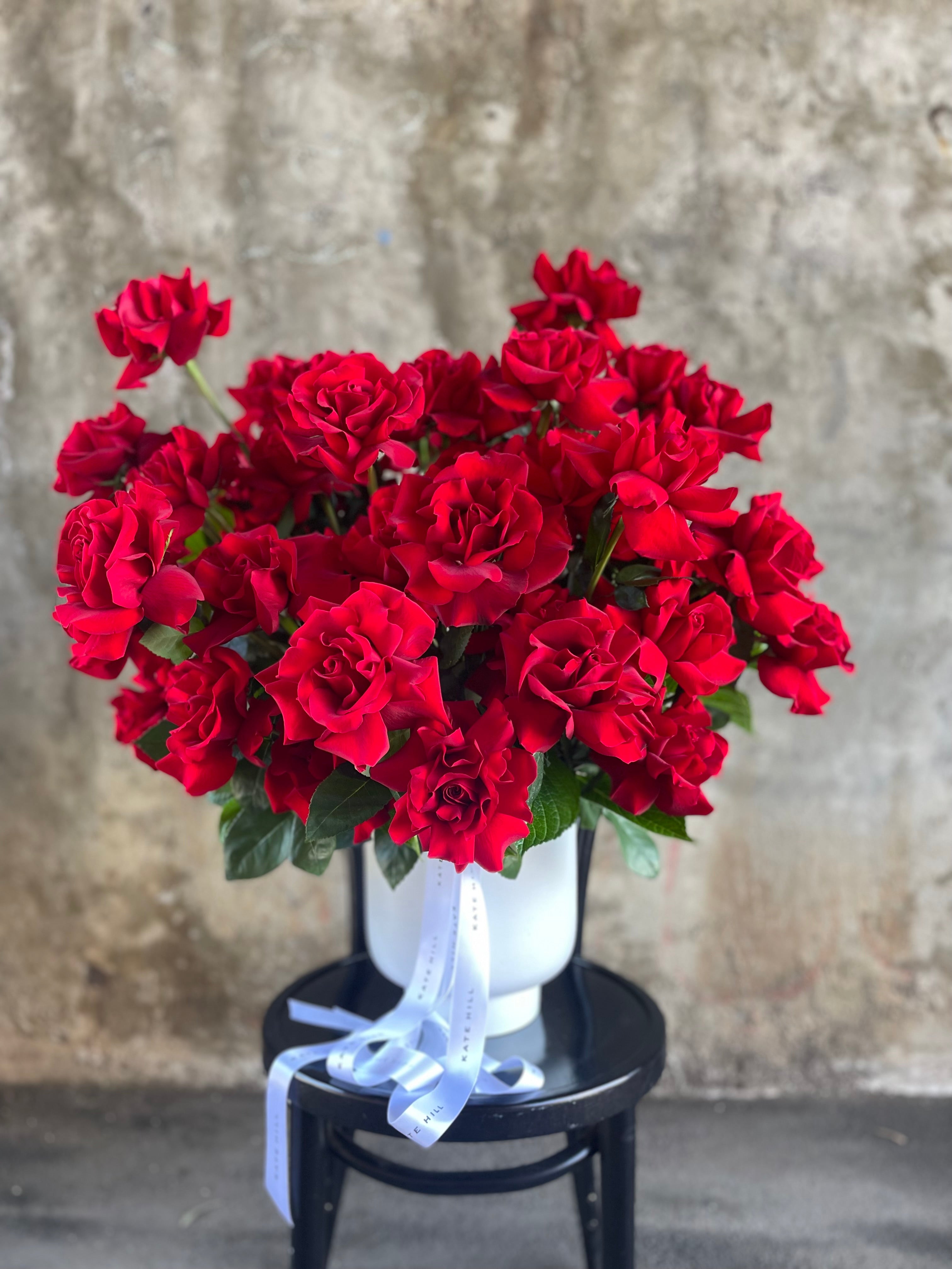 A grande design, displaying 50 long stemmed red roses on mass in a large white footed ceramic vase. Large design is sitting on a black bentwood chair with concrete wall in background.