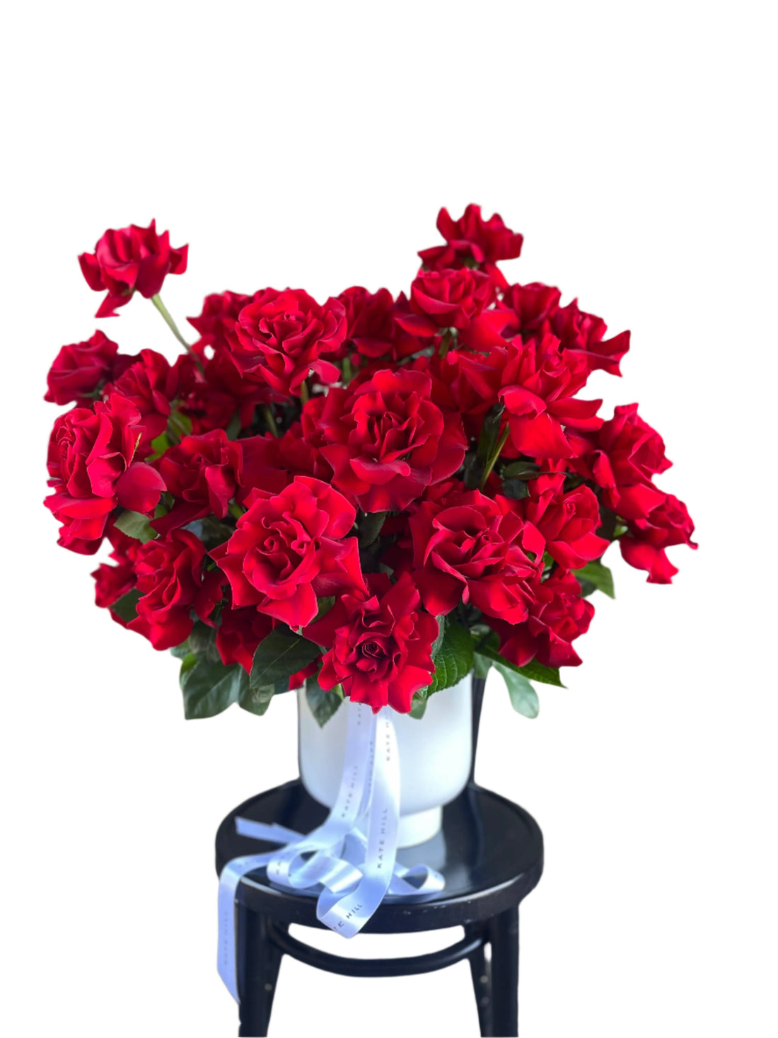 A grande design, displaying 50 long stemmed red roses on mass in a large white footed ceramic vase. Large design is sitting on a black bentwood chair with white wall in background.