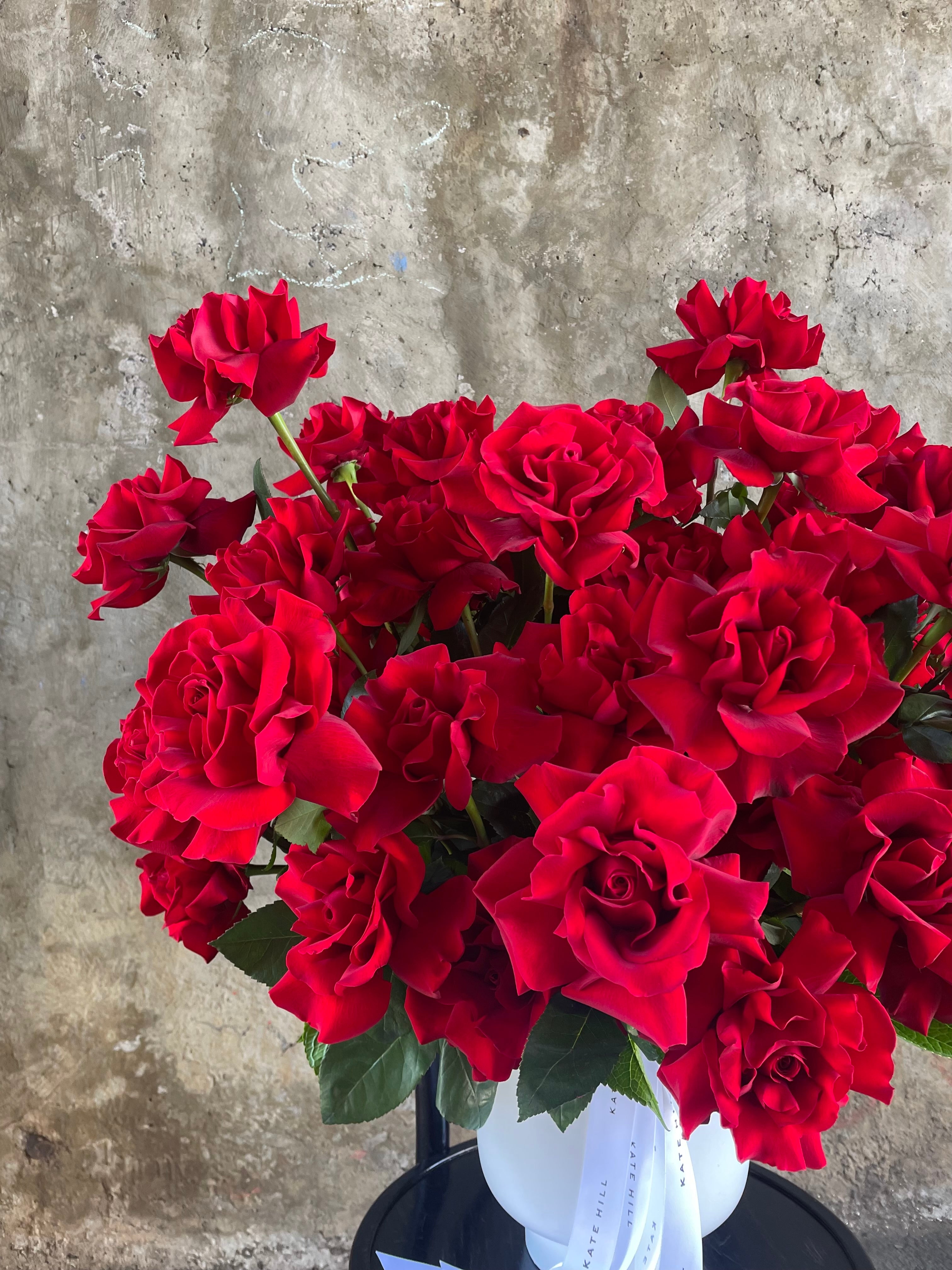 Up close image of A grande design, displaying 50 long stemmed red roses on mass in a large white footed ceramic vase. Large design is sitting on a black bentwood chair with concrete wall in the background.