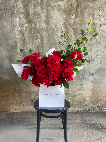 A large red bouquet featuring 24 stems and foliage, wrapped in signature white wrapping and placed into a KHF flower bag. Bouquet bag sitting on a black bentwood chair with concrete wall in background.