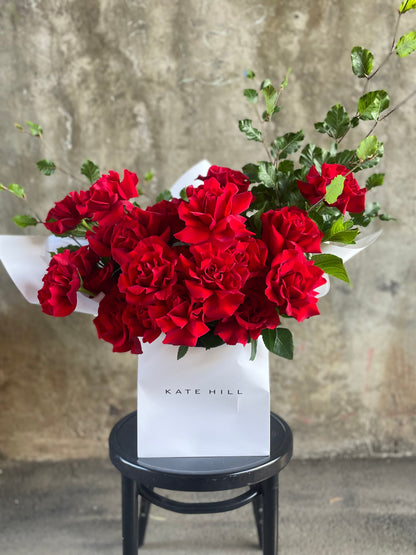 Close up image of 24 red rose bouquet in flower bag. A large red bouquet featuring 24 stems and foliage, wrapped in signature white wrapping and placed into a KHF flower bag. Bouquet bag sitting on a black bentwood chair with concrete wall in background.