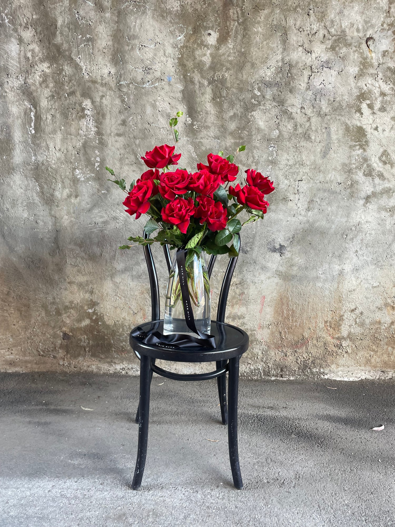 Clear glass vase featuring 10 stems of red roses, sitting on a black bentwood chair in front of a concrete wall.