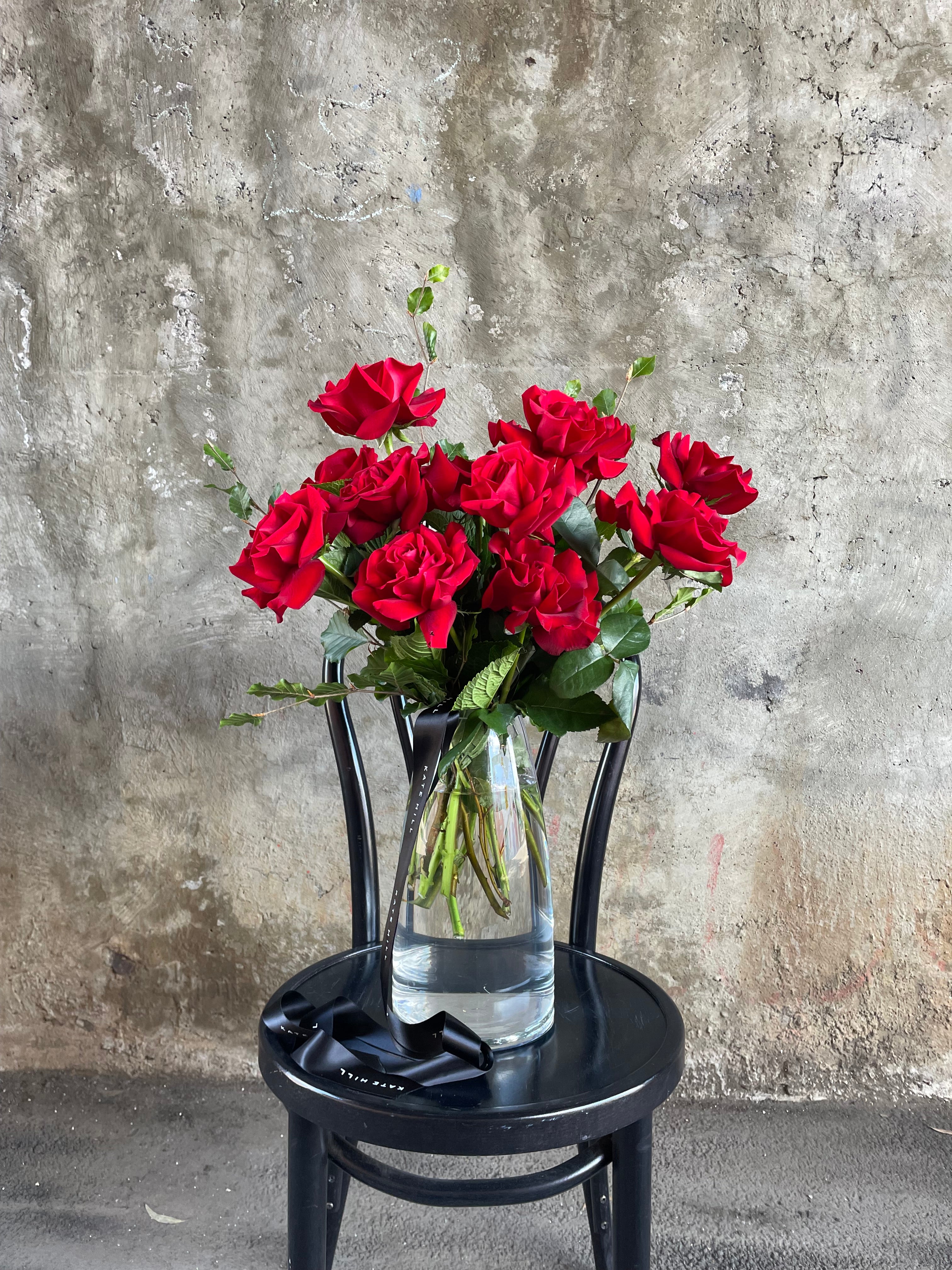 Clear glass vase featuring 10 stems of red roses, sitting on a black bentwood chair in front of a concrete wall.