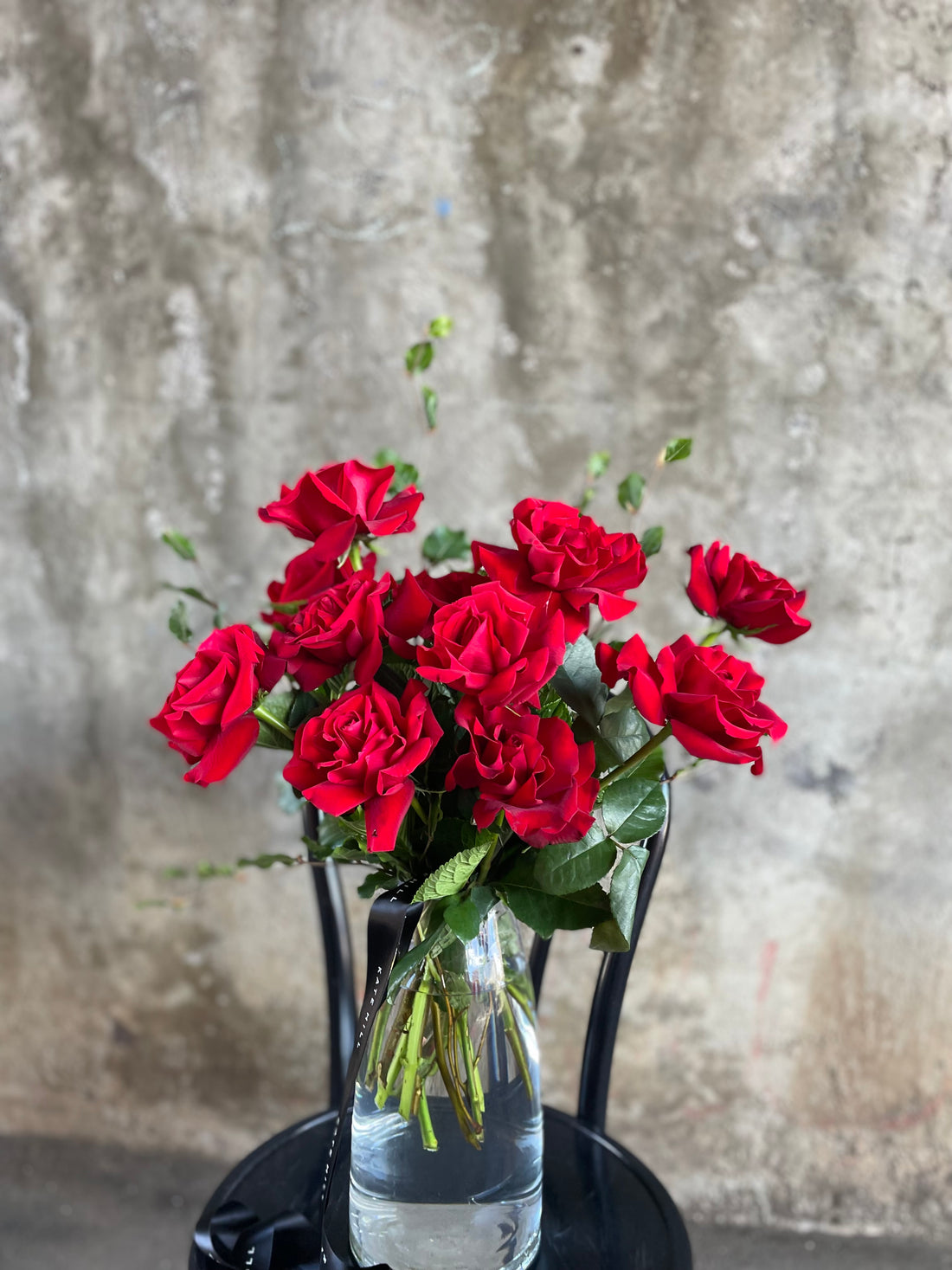Close up image of red roses in clear glass vase. Clear glass vase featuring 10 stems of red roses, sitting on a black bentwood chair in front of a concrete wall.