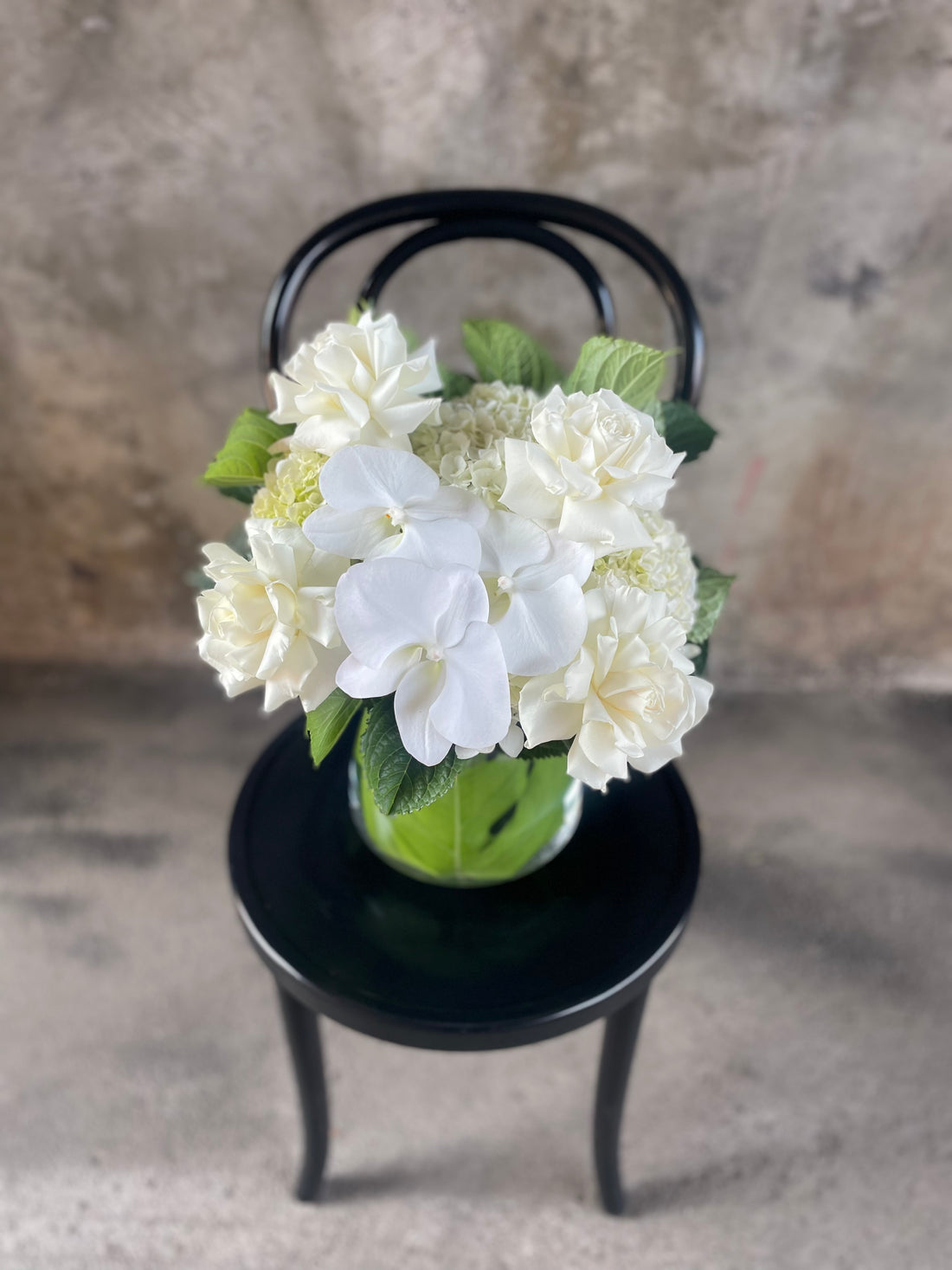 White and green flowers displayed in a glass tapered vase, sitting on a black bentwood chair with a concrete wall background.