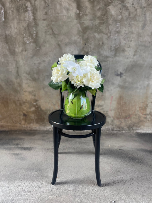 Wide image of White and green flowers displayed in a glass tapered vase, sitting on a black bentwood chair with a concrete wall background.
