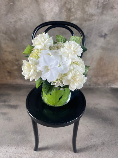 White and green flowers displayed in a glass tapered vase, sitting on a black bentwood chair with a concrete wall background.