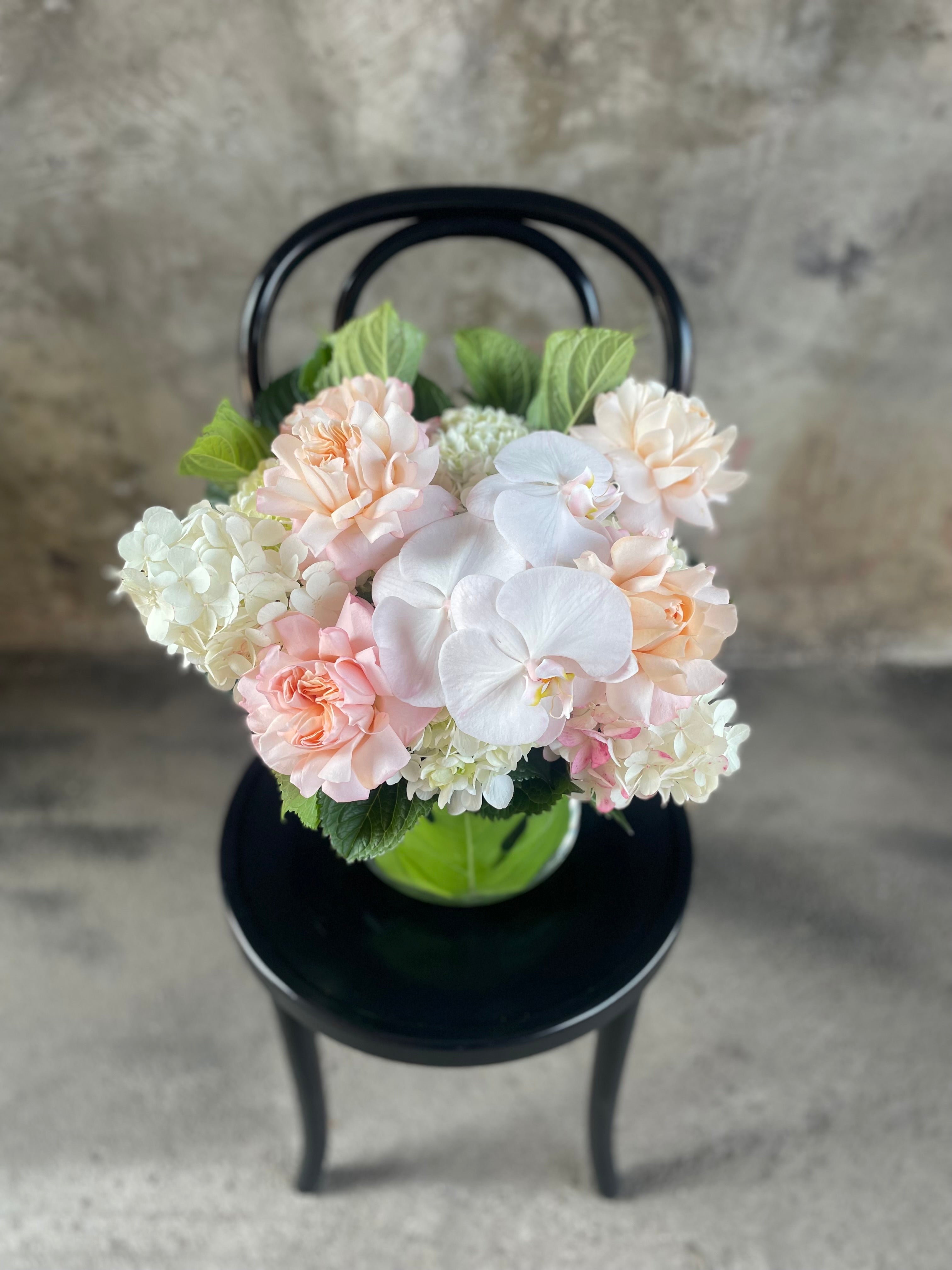 Above angle of Blush white and green flowers displayed in a leaf lined tapered vase, sitting on a black bentwood chair with a concrete wall background.