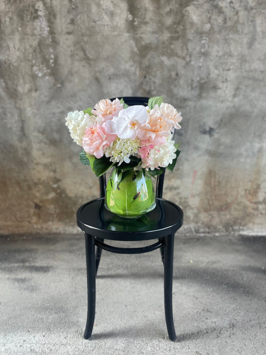 Blush white and green flowers displayed in a leaf lined tapered vase, sitting on a black bentwood chair with a concrete wall background.