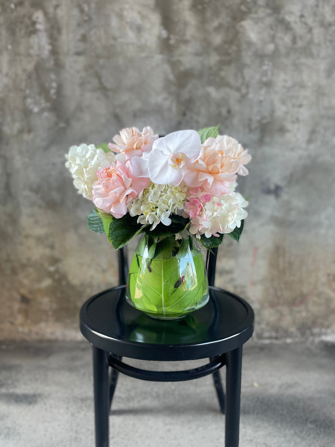 Close up image of Blush white and green flowers displayed in a leaf lined tapered vase, sitting on a black bentwood chair with a concrete wall background.