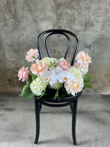 A floral hedge in white, blush and green tones, sitting on a black bentwood chair with a concrete wall background.