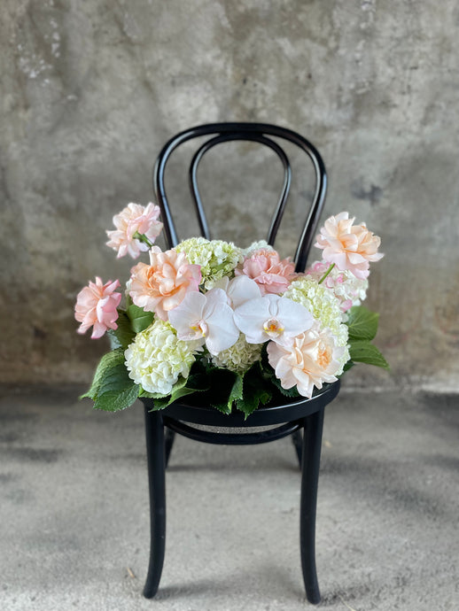 A floral hedge in white, blush and green tones, sitting on a black bentwood chair with a concrete wall background.