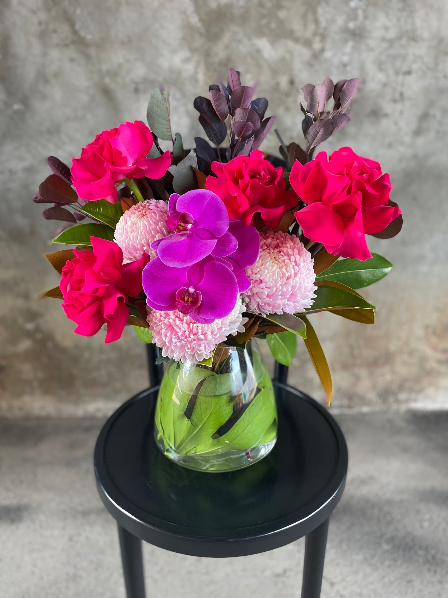 Hot pink, blush, magenta flowers and seasonal foliage displayed in a leaf lined tapered vase, sitting on a black bentwood chair with a concrete wall background.