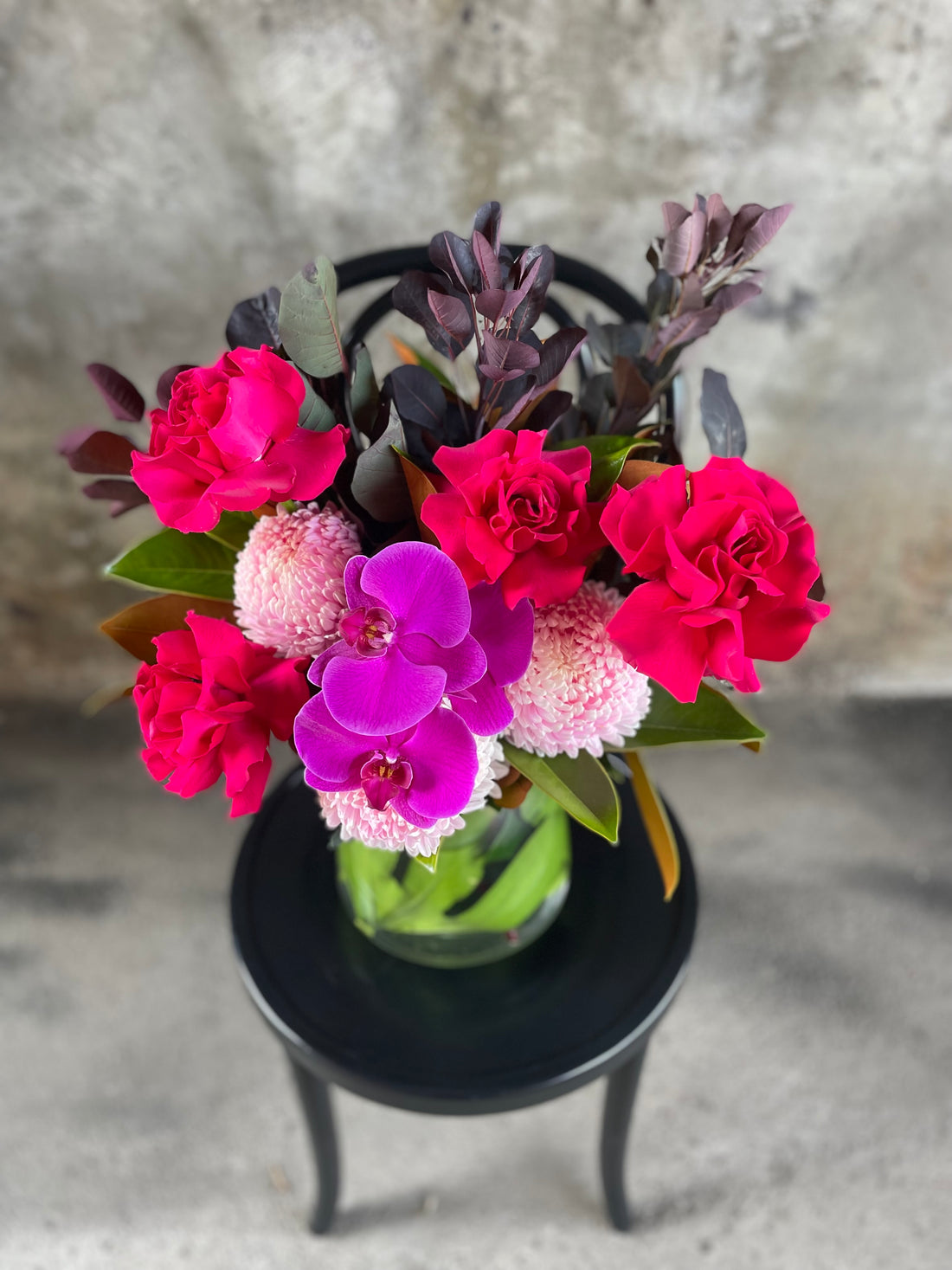 Close up image of Hot pink, blush, magenta flowers and seasonal foliage displayed in a leaf lined tapered vase, sitting on a black bentwood chair with a concrete wall background.