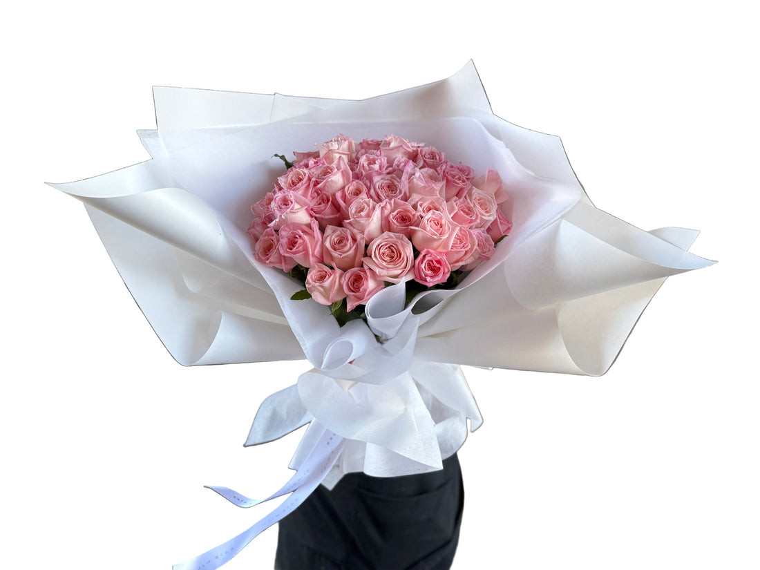 A large and luxe bouquet of pale pink roses wrapped in white paper held against a white wall.