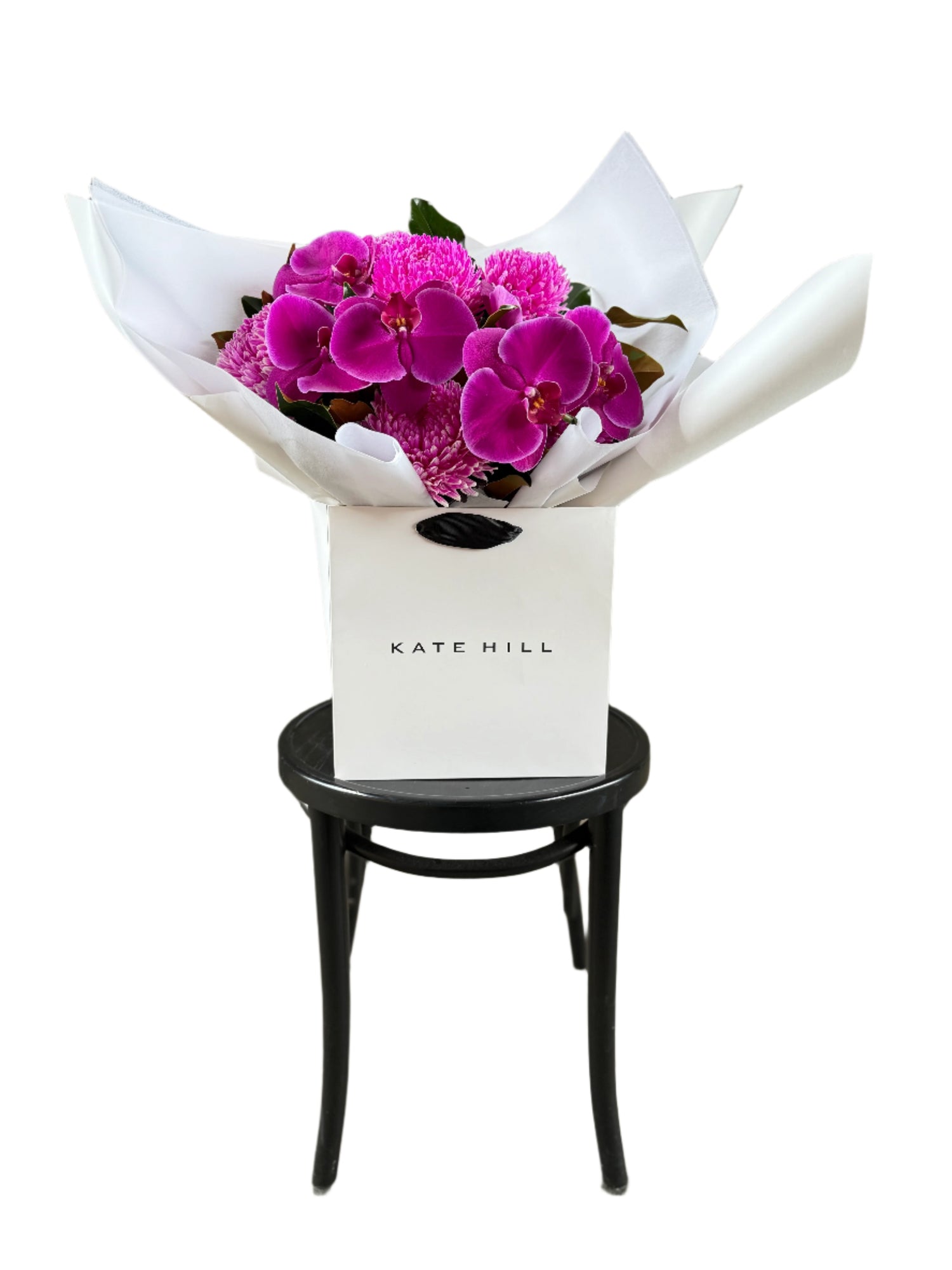 A vibrant hot pink and magenta flower bouquet, presented in our signature white wrapping and flower bag. Bouquet bag is sitting on a black bentwood chair with a white background.