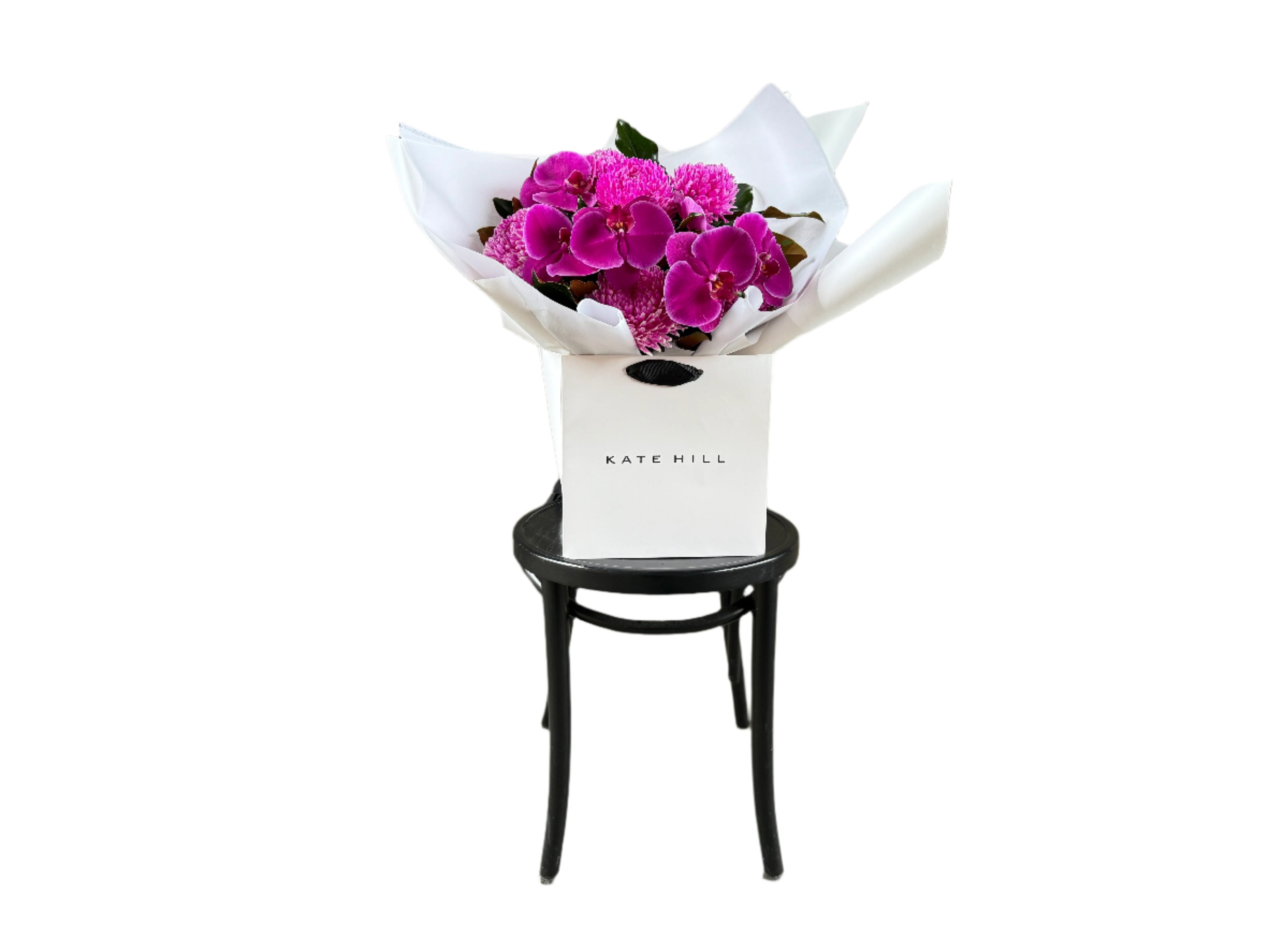 Wide image of a vibrant hot pink and magenta flower bouquet, presented in our signature white wrapping and flower bag. Bouquet bag is sitting on a black bentwood chair with a white background.