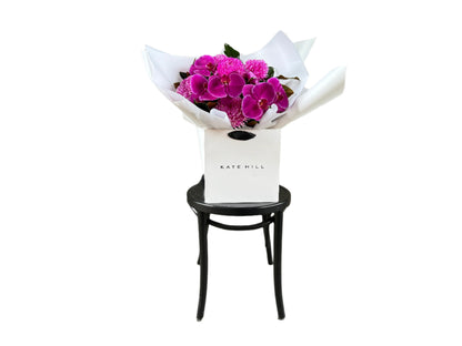 Wide image of a vibrant hot pink and magenta flower bouquet, presented in our signature white wrapping and flower bag. Bouquet bag is sitting on a black bentwood chair with a white background.