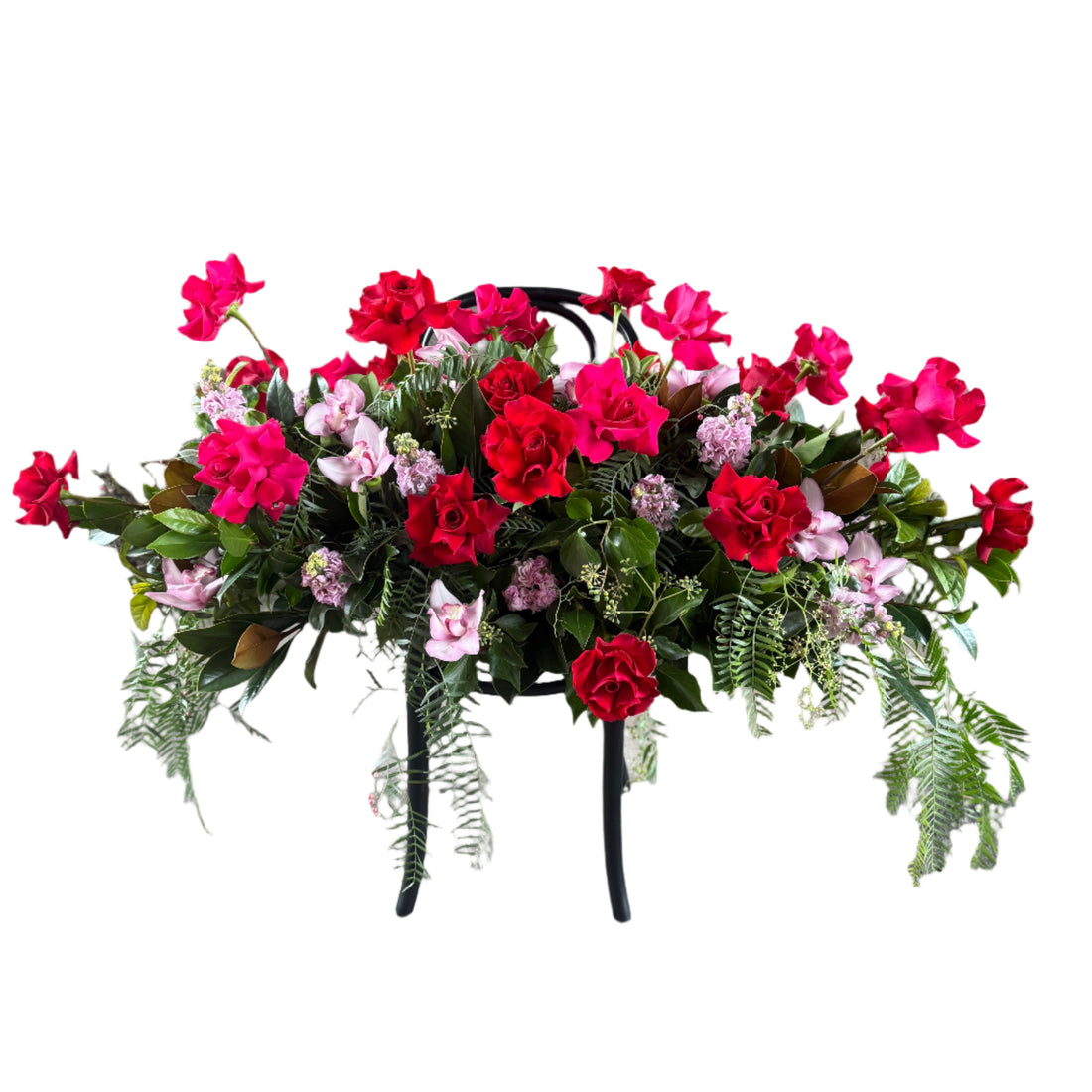 A funeral casket spray displaying hot pink roses, pale pink seasonal flowers and green seasonal foliages. Casket spray sitting on black bentwood chair with white background.