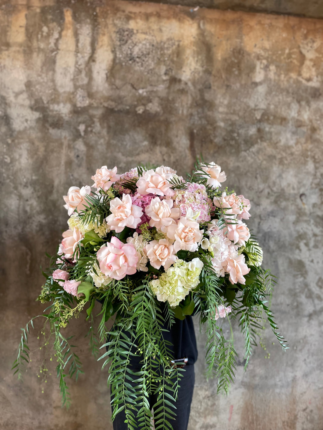 Pastel pink, white and green casket design, held by a florist wearing back, up against a concrete wall.
