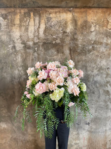 Wide image of a Pastel pink, white and green casket design, held by a florist wearing back, up against a concrete wall.