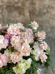 Classic blush roses, blush hydrangeas, lilac stock and foliages feature in a Pastel pink, white and green casket design, held by a florist wearing back, up against a concrete wall.