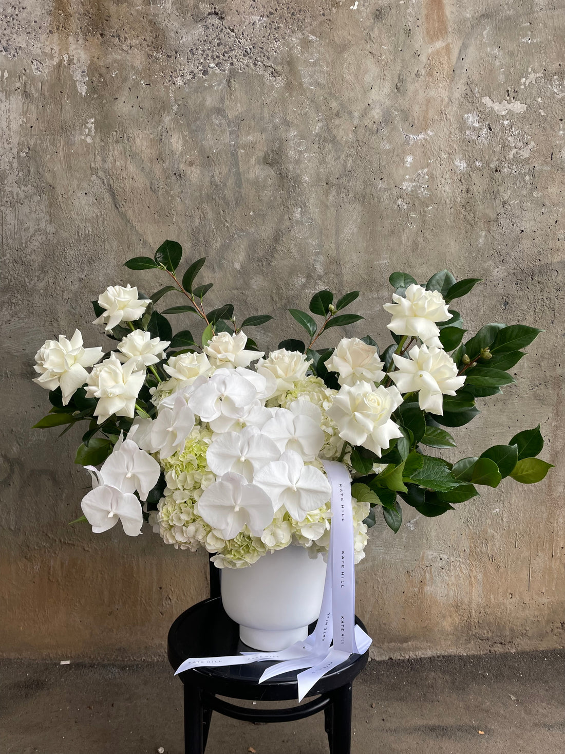 A large luxe white flower design sitting on a black bentwood chair with concrete wall behind.